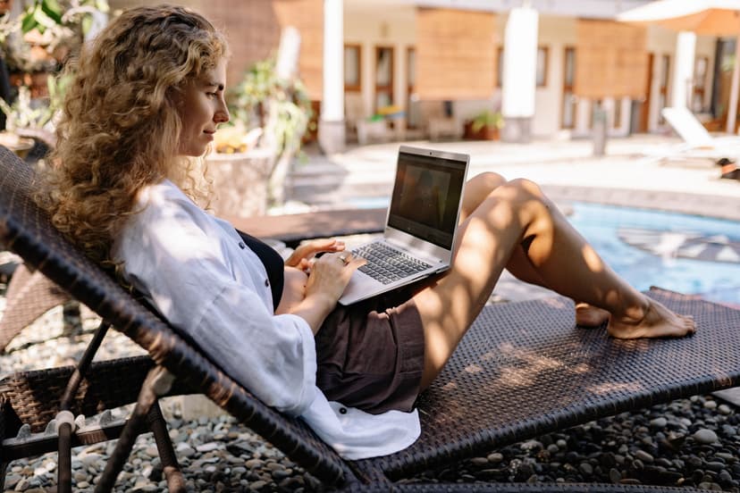 Una mujer trabaja con su laptop recostada en una tumbona junto a una piscina
