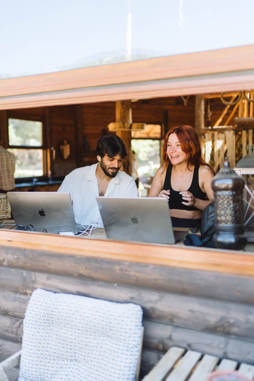 Dos personas trabajan con sus laptops en la terraza de una cabaña de madera, riendo