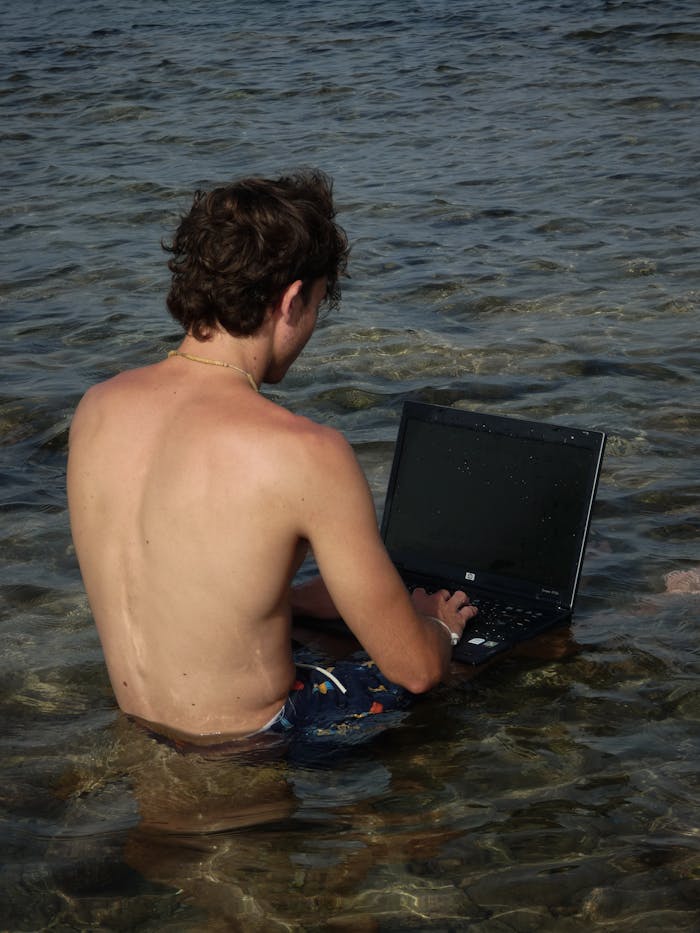 A shirtless man sits in shallow coastal water, using a laptop, embodying the digital nomad lifestyle.