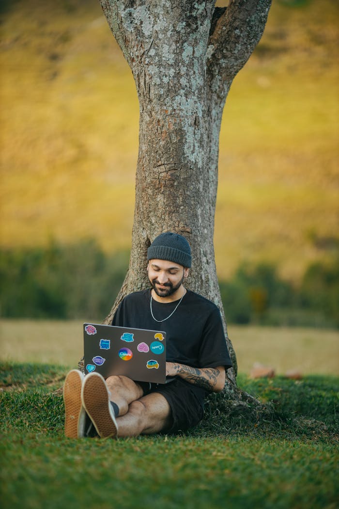 A man with a laptop sits smiling against a tree in a rural meadow, enjoying a peaceful moment outdoors.