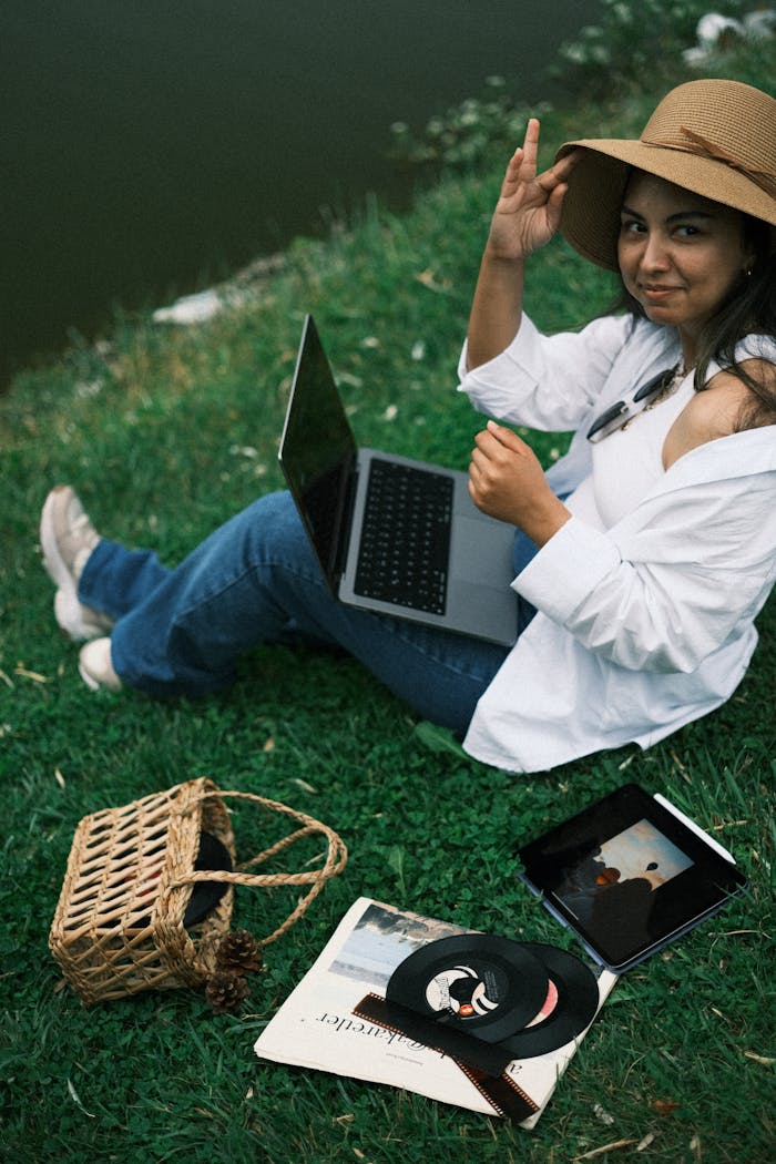 A woman using a laptop and tablet while sitting on grass by a lake in summer.