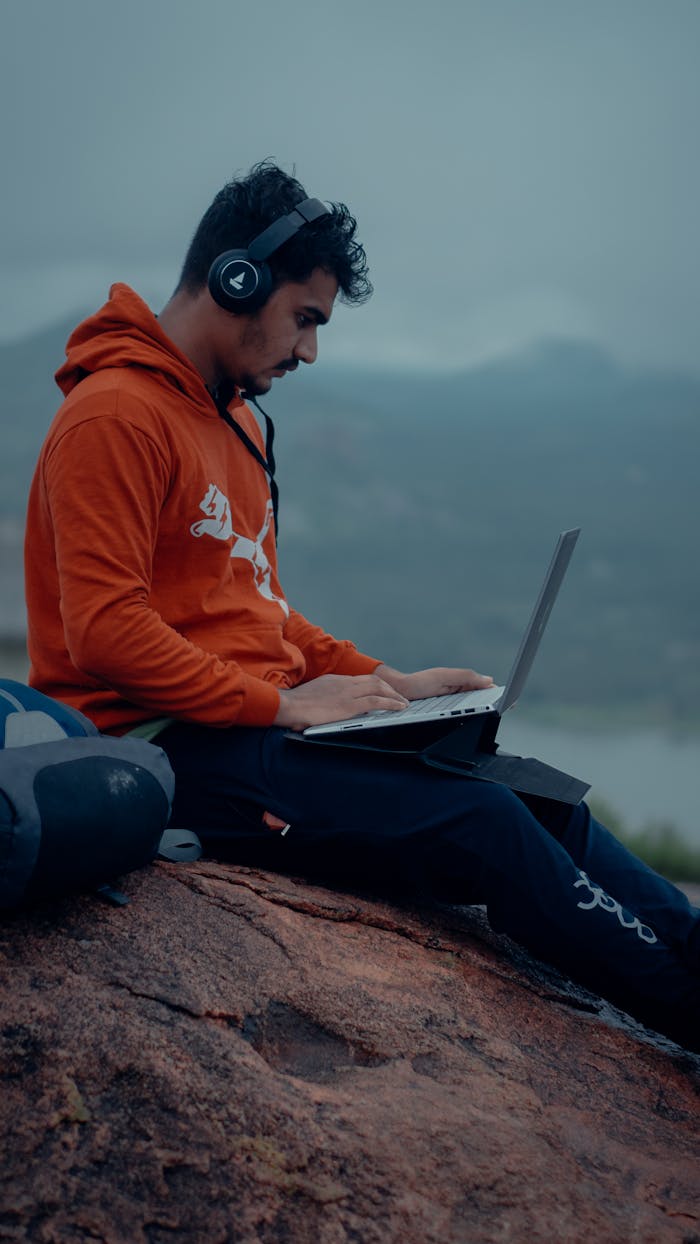 Man in hoodie using laptop outdoors with headphones, capturing modern remote work.